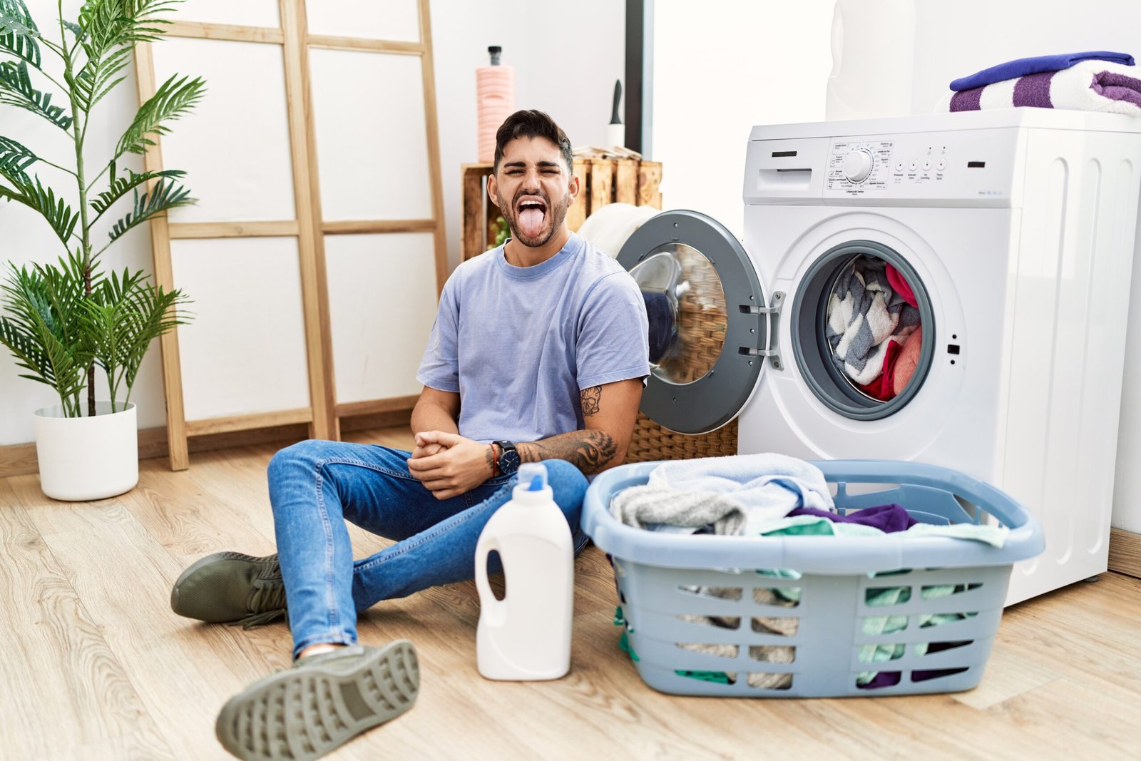 young hispanic man putting dirty laundry into washing machine sticking tongue out happy with funny expression. emotion concept.